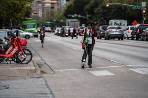 En gata i Austin i Texas. I förgrunden till vänster parkerade elsparkcyklar. På gatan står en person med sin sparkcykel. I bakgrunden en kvinna på en sparkcykel, bilar och människor.