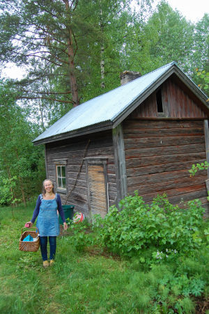 Selma Kilpi seisoo hymyillen vanhan saunan edessä kädessään kori.