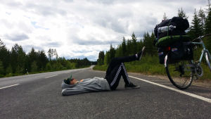 Matti makaa moottoritiellä kädet päänsä alla ja toinen jalka ylhäällä pakattu pyörä vierellään.