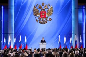 Putin talar om läget i nationen. En stor blå bakgrund med en dubbelörn och en man i en talarstol omgiven av ryska flaggor. I förgrunden huvuden tillhörande publiken. President Vladimir Putin talar om läget i nationen.
