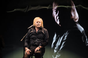 Mustaan paitaan pukeutunut Danny istuu lavalla jakkaralla mikkistandin edessä. Taustascreenillä Danny itse.
