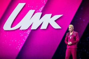 Magentan väriseen pukuun ja glittervaljaisiin pukeutunut Antti Tuisku seisoo lavallla screenin edessä, jossa lukee UMK.