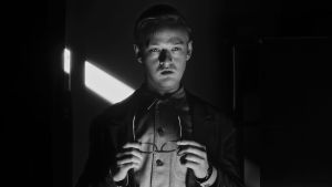 Nuori mies seisoo silmälasit kädessään mustavalkoisessa kuvassa.