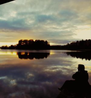 Mies istuu iltahämärässä järven rannalla, vedestä heijastuvat pilvet ja auringonlaskun kajo ja saari kauempana.