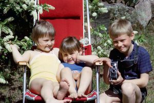 Sisarukset Marikki, Riikka ja Jyrki Hakola Raumalla mummolan mökillä kesällä 1965. Marikki kainalossaan Riikka istuu kesäisellä pihalla aurinkotuolilla, veli Jyrki on kumartunut kameran kanssa heidän viereensä.