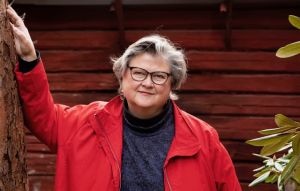 Marikki Hakola MArikki HAkola seisoo hirsirakennuksen punamultaisen seinän edessä punainen takki päällään.
