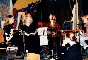 Heristä kukkaa -yhtye jyväskyläläisen opiskelijaravintola Ilokiven yläkerrassa keikalla vuonna 1992. Bassoa soittaa Niina (vas.), lapsuudenystävä Minna Malja ja hänen vieressään kitara sylissään Anita Seppä. Oikeassa reunassa kitaraa soittaa Matti. Opiskelijoiden bändi soittamassa tuoleilla istuen. Kuvassa viisi soittajaa sekä vahvistimia, nuottitelineitä ja mikrofonitelineitä.
