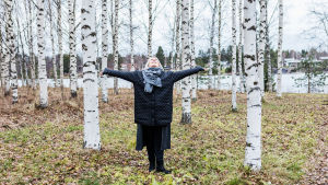 Elina Koskelainen seisoo koivujen keskellä kädet levällään ja katse ylös puiden latvaan.