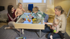 Hanna (till höger) med dottern Bella i famnen sköter om syster Sanjas två barn tjugo timmar i veckan. Till vänster i bilden sitter en kvinna som håller en flicka som spelar Kimble med sin äldre bror. Till höger en annan kvinna med en liten flicka i famnen.