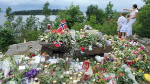 Utsikt över Utøya 2011. Rosor, flaggor och ljud på en stor sten, en vuxen med två barn pekar över fjorden mot Utöya