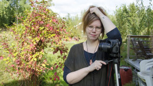 Alexandra fotar nästan alltid bilderna själv, med hjälp av kamerastativ och fjärrkontroll. En kvinna står och lutar sig mot en kamera som står på ett stativ. Hon står i en trädgård. Det är sensommar.