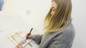 Caroline satsar gärna lite extra på sina pennor. Caroline Eriksson har långt blont hår och en grå tröja. Hon lutar sig över ett bord med ett häfte framför sig. I handen håller hon en penna och hon ritar en bokhylla i häftet.