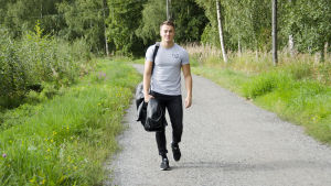 En man iklädd svarta jeans och en grå t-skjorta går på en grusväg kantad med grönt gräs och blommor. Han ler och ser glad ut.