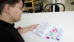 Zarah har ritat av ett gammalt foto på hen och hens bror. Zarah Holmberg tittar i tidningen hen gjort. I tidningen finns en färgglad teckning på hen och hens bror.
