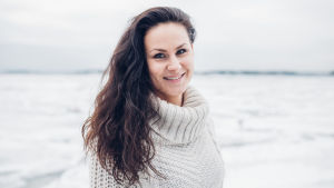 Elina Hovinen seisoo talvisessa maisemassa villapaidassa ja hymyilee kameralle.
