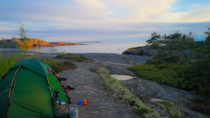 Många finländare har vistats i skärgården i år. Ett grönt tält har spänts upp på en skärgårdsö. Solen håller på att gå ner över viken, där kajaker är uppdragna på klippstranden.