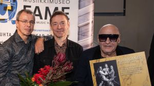 Remu Aaltonen (höger), Ile Kallio och Janne Louhivuori tog emot ett diplom då Hurriganes blev vald till Finnish Music Hall of Fame torsdagen 19.9.2019. Remu Aaltonen, Ile kallio och Janne Louhivuori poserar med en blombukett och med en Hurriganes-plansch. Remu har solglasögon på sig.