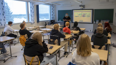 En lärare med munskydd står framför ett klassrum fullt av elever.