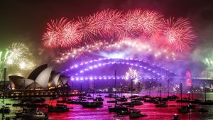 Fjolårets fyrverkerishow vid tolvslaget i Sydney, Australien var i vanlig ordning spektakulärt. Även i år vankas det fyrverkerier men strategiska utsiktsplatser har stängts av för att undvika folksamlingar – trots relativt få fall av covid-19. Vy över hamnen i Sydney, med en bro i mitten och operahuset till vänster, med röda fyrverkerier som lyser upp himlen.