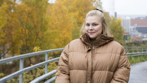 Julia Andersson kallar sig gärna tjock - det är inte något negativt. En blond kvinna iklädd en tjock beige dunjacka. Hon ler. I bakgrunden syns träd med gula höstlöv.