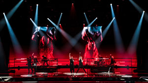 Blind Channel -yhtye lavalla punaisen screenin edessä keskisormet pystyssä. Taustacreenillä laulajat roikkuvat ylösalaisin.