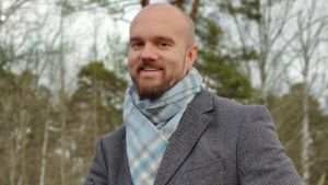Jouni Heiskanen är ny direktför för Tvärminne zoologiska station och två andra forskningsstationer. Han doktorerade 2015 vid bio- och miljövetenskapliga fakulteten. Förenklat kan man säga att hans forskning handlar om klimatförändring och vattendrag. En skäggig man med rakat huvud, klädd i grå kavaj och ljusblå halsduk sitter på en sten. I bakgrunden nakna björkar och knotiga tallar.
