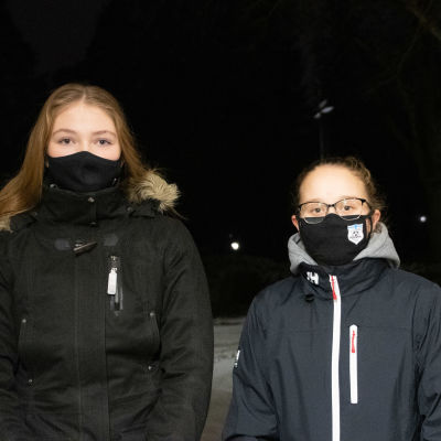 Två flickor står i mörker med munskydd på sid.