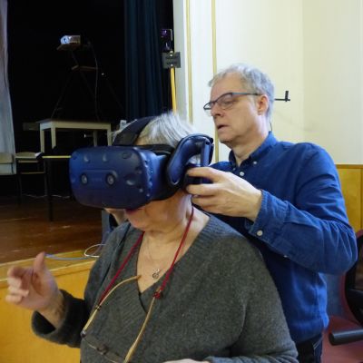 En kvinna sitter i en stol medan en man klär på henne ett par VR-glasögon.