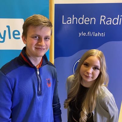 Pitkä nuori mies sinisessä paidassa ja vieressä häntä lyhyempi vaalea, pitkähiuksinen nuori nainen. Molemmat hymyilevät, taustalla näkyy logot Yle ja Lahden Radio. 