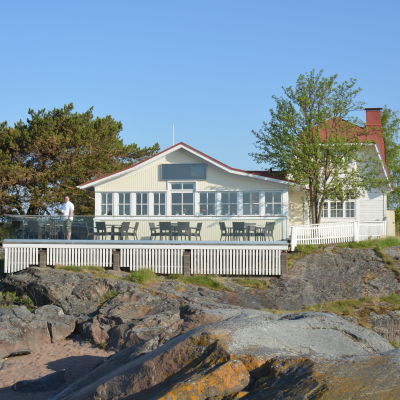 En gammal vit trävilla med rött tak vid havet, på uteserveringen står en man.