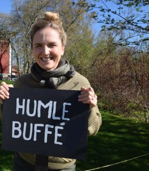 Framför ett rött hus står en leende kvinna med en skylt "Humle Buffet"