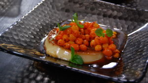En stor skopa havtorn bryter sötman i Pauls crème caramel. En portion av efterrätten creme caramel toppad med rårörda havtorn och några citronmelissblad. Efterrätten är serverad på ett grått, fyrkantigt glasfat.