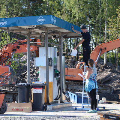 En ny bränslestation installeras på ett byggområde. Två personer står och inspekterar stationen. I bakgrunden syns en orange grävskopa.