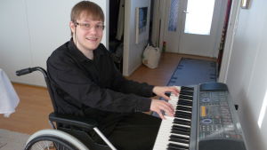 Tommy vid sitt piano. Tommy Lundström sitter i rullstol, spelar piano och tittar mot kameran