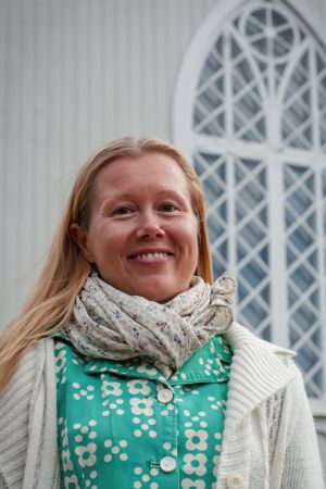 Sini Tammisalo guidade i kyrkan på sommaren 2018. Vaaleatukkainen nainen huivi kaulassa, taustalla vaaleaa seinää ja koristeellinen kirkon ikkuna