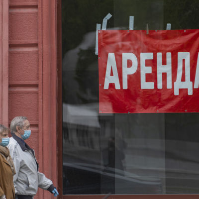Man och kvinna som bär munskydd promenerar på gata vid skyltfönster med röd skylt där det står ''uthyres'' på ryska.