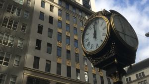 Klocka vid Trump Tower i New York. Klockan vid Trump Tower visar någon minut över 12. I bakgrunden skyskrapa och moln.