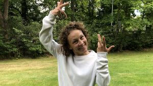 Natasha Lommi dansar rollen som Covid i Nationaloperans nygamla opera Covid fan tutte. Kvinna med lockigt hår och vit blus vid en gräsmatta dansar.
