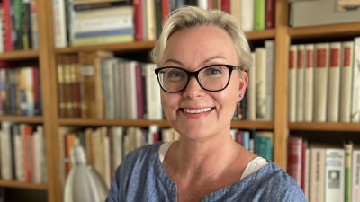 Nelly Laitinen håller i en hög med böcker framför sin bokhylla.