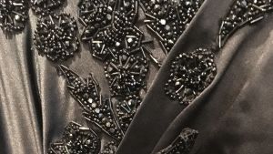 En detalj av en svart sidenklänning broderad med svarta glaspärlor. Ett svart tyg med dekorationer av svarta glaspärlor.