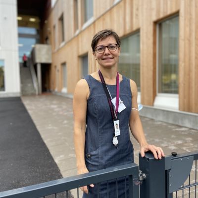 En kvinna står bakom ett lågt staket, med en modern byggnad i bakgrunden. Hon ser glad ut.