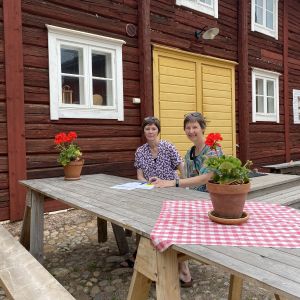 Susanne Lagus och Gunilla Sand sitter utomhus vid ett bord med papper framför sig på bordet och en röd stockvägg bakom sig.