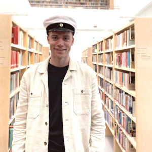 Personporträtt på ung man, han har studentmössa på huvudet, bär ljus jacka och ler. I bakgrunden bokhyllor i biblioteksmiljö.