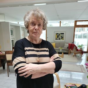 En äldre dam med grå lockar står med armarna i kors och tittar med bestämd bild i kameran