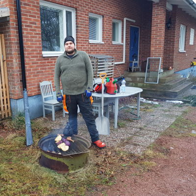 Viktor Eriksson stor framför sitt hus, bredvid en eld där han lagar mat.