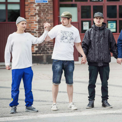 Fem unga personer klädda i hiphop-kläder håller varandra i händerna framför ett gammalt lokstall.