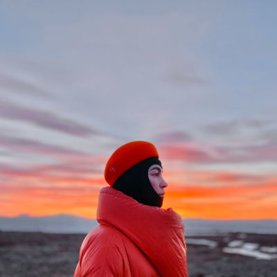 Meeri Koutaniemi katsoo kuvan oikean reunaan. Hän seisoo lähellä kameraa punainen paksu takki ja punainen hattu päässään. Taivaalla auringonnousu.
