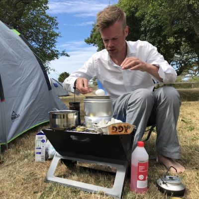En man i vit skjorta kokar ägg på ett spritkök utanför sitt tält.