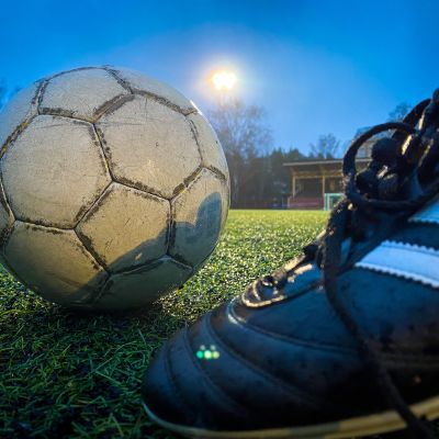 Jalkapallokenkä ja jalkapallo kuvattuna Pyynikin tekonurmikentällä. Takana kajastaa kentän valonheittimen valo.
