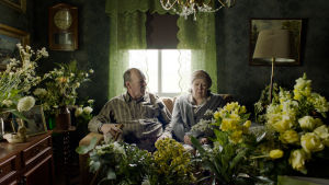 "Jag vill ju bara hjälpa!" Elsa (Lena Labart) förstår inte alls varför brodern Nisse (Peik Stenberg) blir irriterad då hon möblerar om. Nisse (Peik Stenberg) och hans syster Elsa (Lena Labart) sitter i en soffa i ett rum fullt med blombuketter.